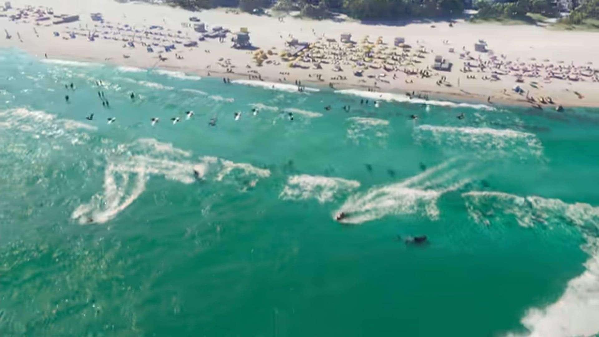 Jet skis and swimmers in turquoise water off a beach lined with umbrellas, seen from above