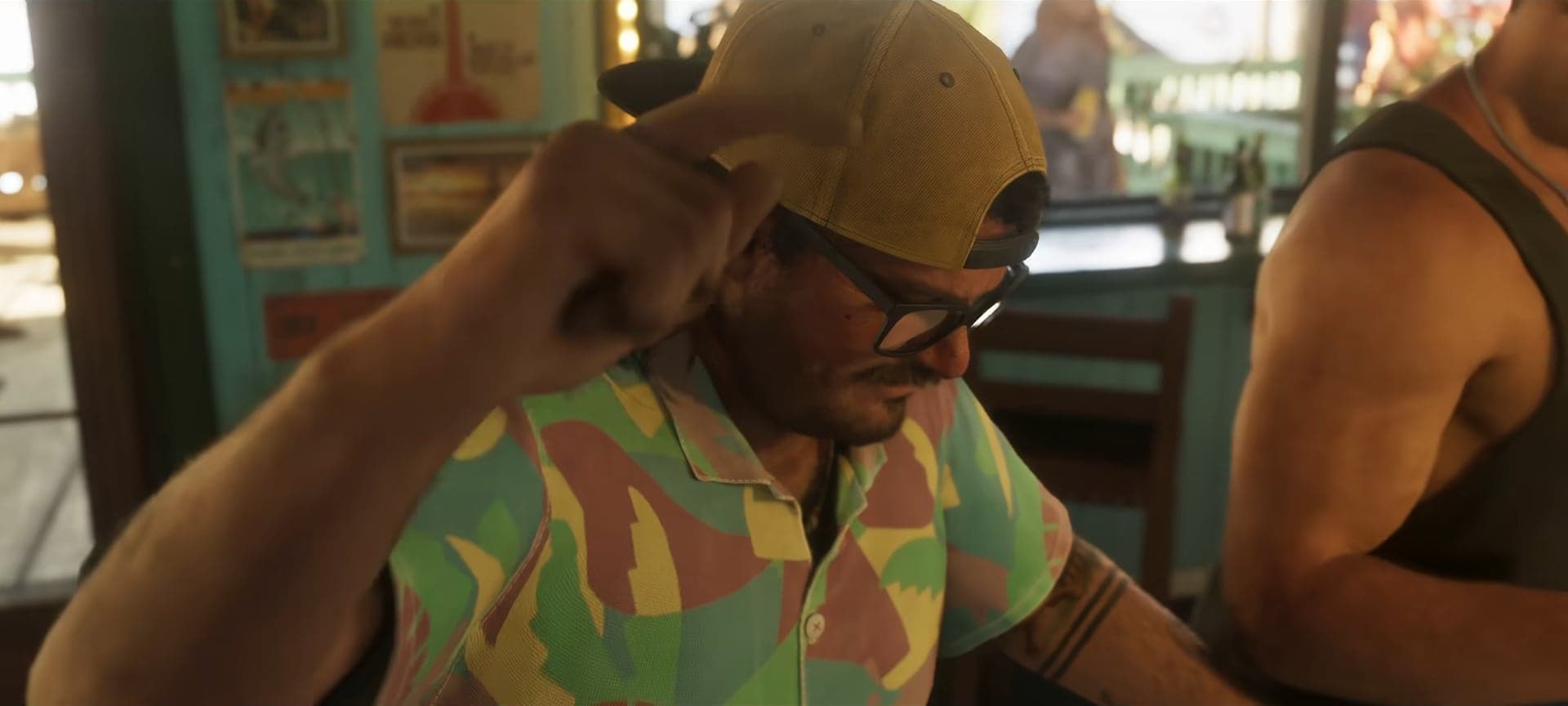 A man in a yellow cap, glasses and a patterned shirt raising his hand in a bar